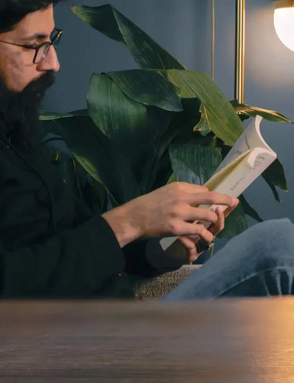 A bearded guy wearing glasses, a black shirt and jeans; sitting and reading a book with a green plant and a blue wall in the background and a blurred table in the foreground.