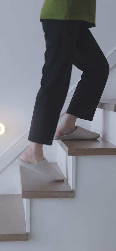 A side view of girl wearing dark pyjamas and fuzzy warm slipper walking up a brown wooden stairs and a light on the wall near the stairs shining