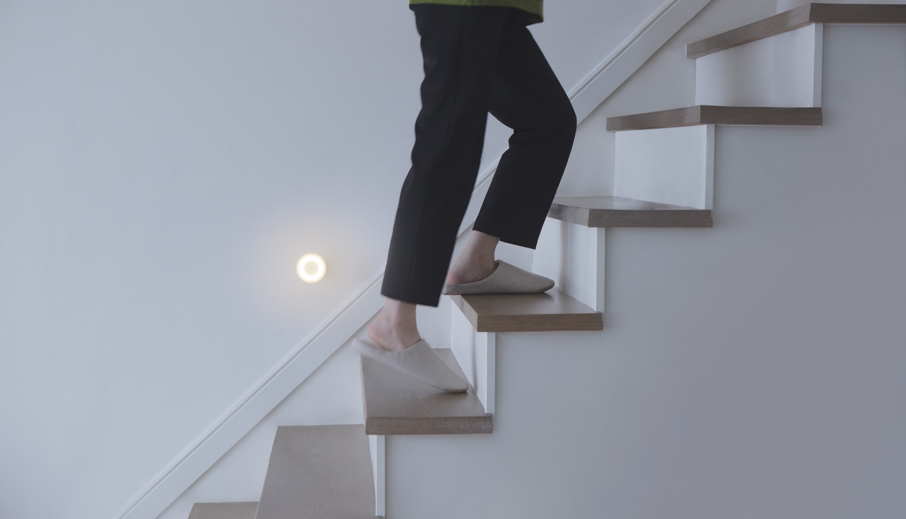 A side view of girl wearing dark pyjamas and fuzzy warm slipper walking up a brown wooden stairs and a light on the wall near the stairs shining